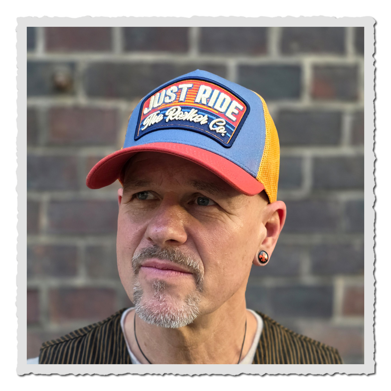 Mann trägt vor Mauer Rokker Cap Just Ride – Trucker Cap in Blau, Rot und Gelb